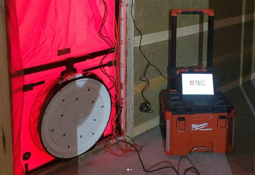 A blower door setup installed in a home's front doorway, with a red canvas and a large circular fan at the bottom. An attached control panel and laptop are visible on the right side, used to measure the building's air leakage